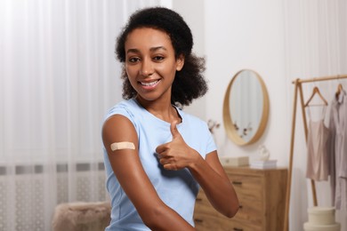 Photo of Happy young woman with adhesive bandage on her arm after vaccination showing thumb up indoors