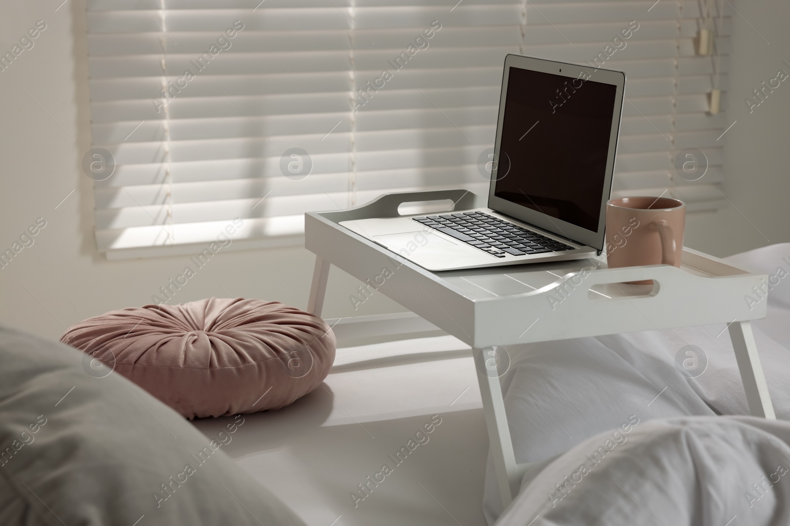 Photo of White tray table with laptop and cup of drink on bed indoors