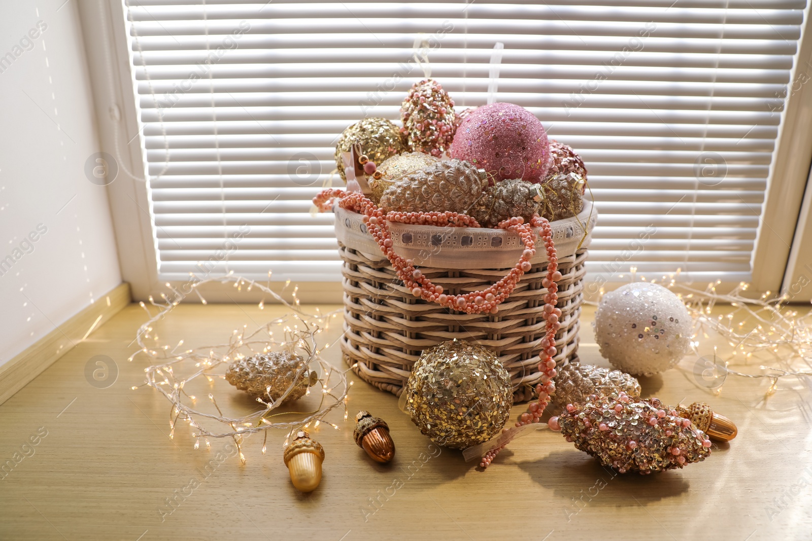 Photo of Basket with beautiful Christmas tree baubles and fairy lights on window sill indoors