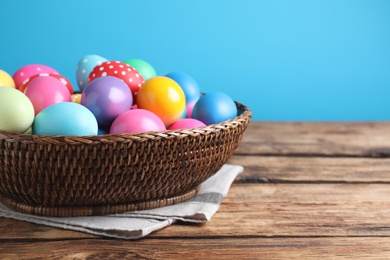 Colorful Easter eggs in wicker basket on wooden table against light blue background, closeup. Space for text