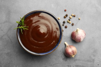 Tasty barbeque sauce in bowl, rosemary, garlic and peppercorns on grey textured table, flat lay