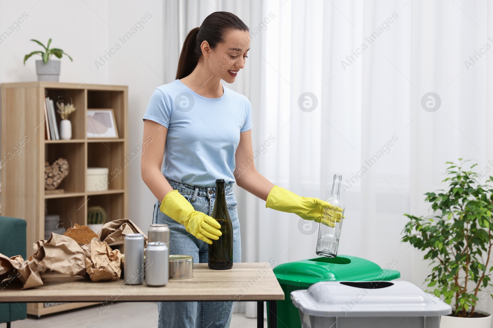 Photo of Garbage sorting. Smiling woman throwing glass bottle into trash bin in room