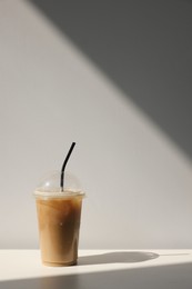 Photo of Plastic takeaway cup of delicious iced coffee on white table under sunlight, space for text