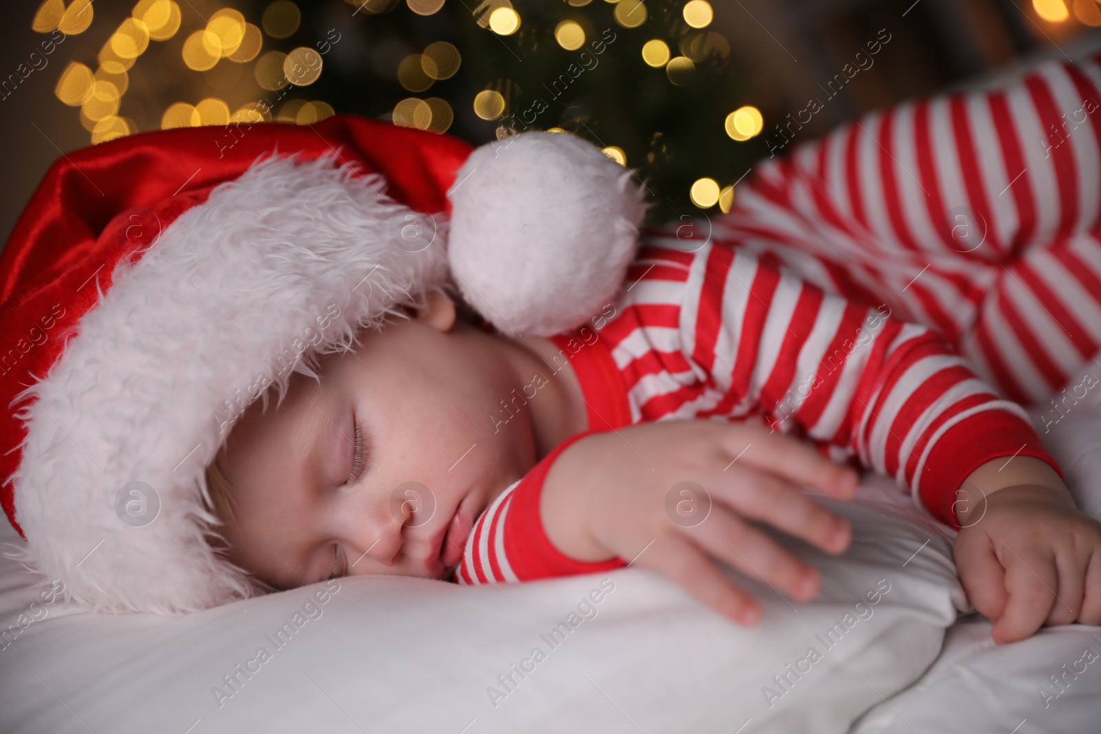 Photo of Baby in Christmas pajamas and Santa hat sleeping on bed indoors