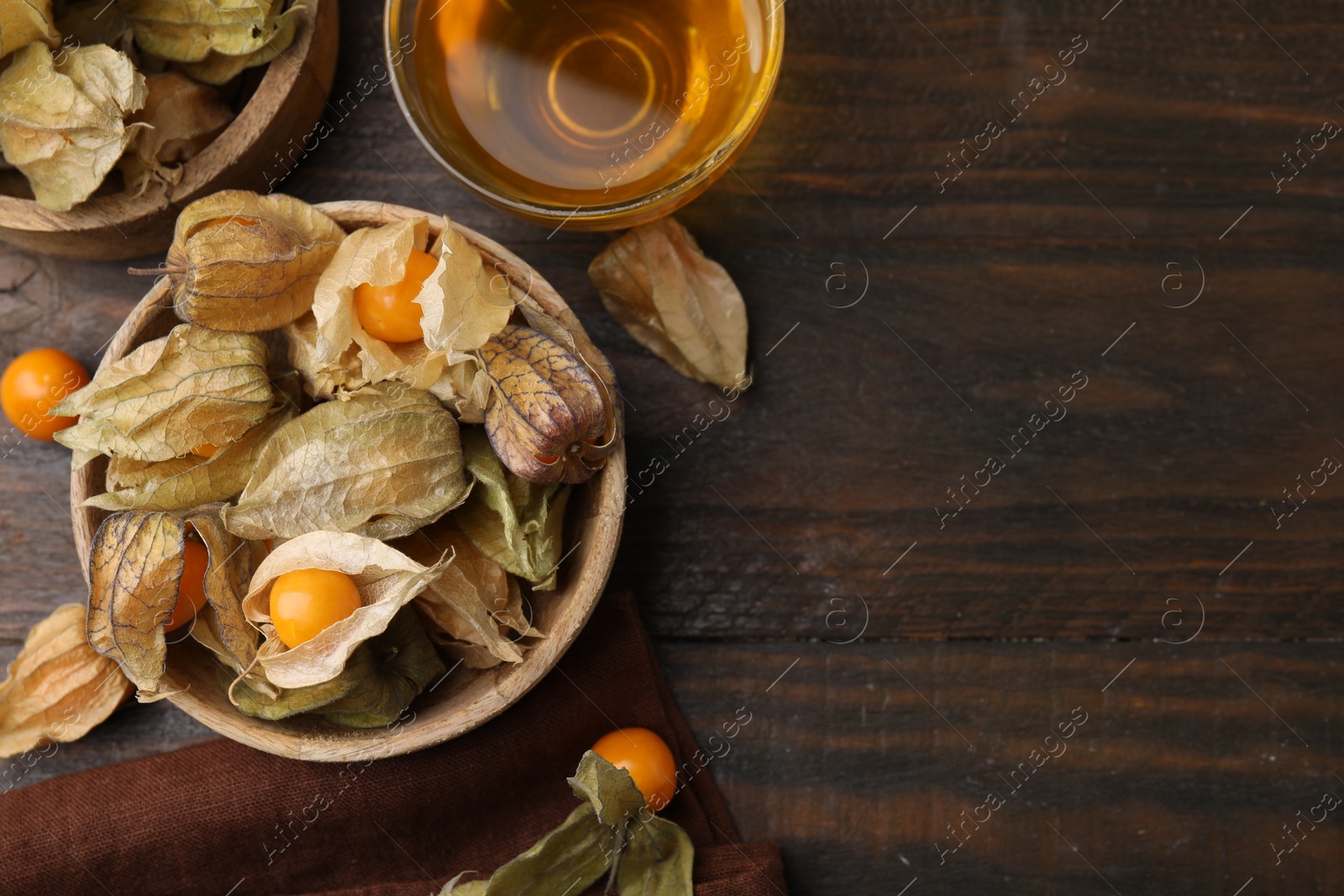 Photo of Ripe physalis fruits with calyxes in bowls and cup of tea on wooden table, flat lay. Space for text