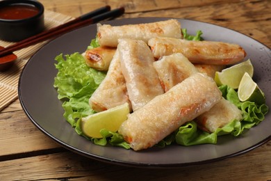 Photo of Plate with tasty fried spring rolls, lettuce, lime and chopsticks on wooden table, closeup