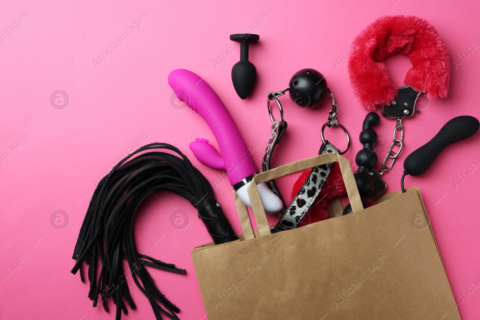 Photo of Paper shopping bag with different sex toys on pink background, flat lay