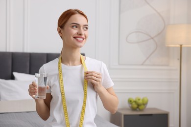 Photo of Happy young woman with measuring tape and glass of water in room, space for text. Weight loss