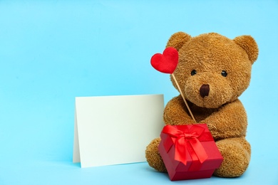Photo of Cute teddy bear with red heart, gift box and blank card on light blue background. Valentine's day celebration