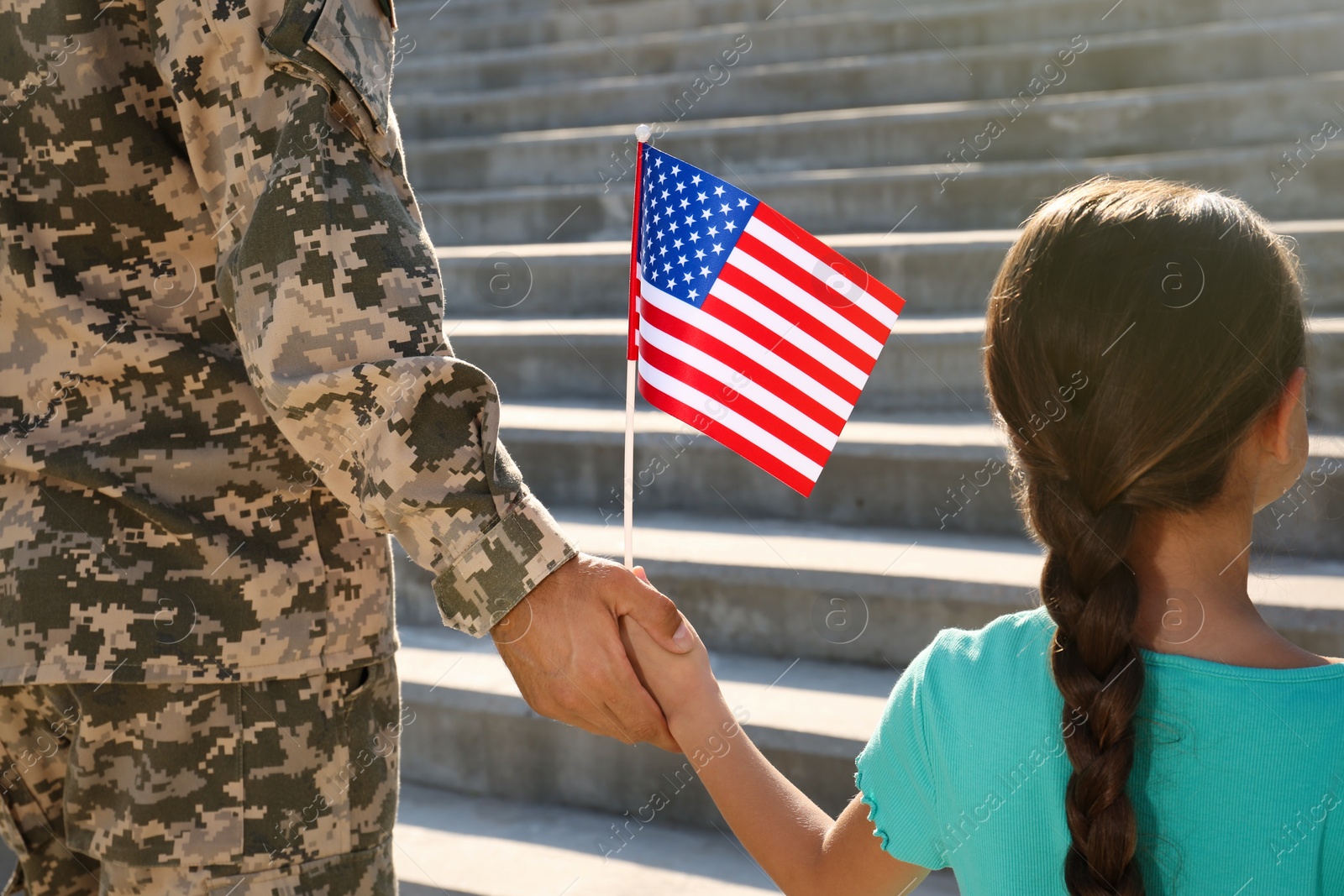 Photo of Soldier and his little daughter with American flag holding hands outdoors, back view. Veterans Day in USA