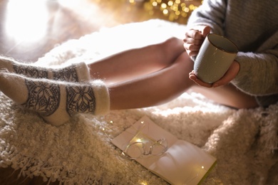 Photo of Woman with cup of hot beverage and book at home in winter evening, closeup