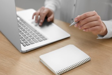 Woman with notebook and pen working on laptop at wooden table, closeup. Electronic document management