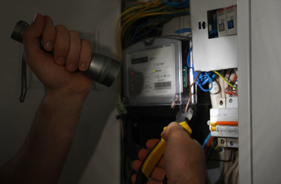 Electrician with flashlight fixing electric panel indoors, closeup