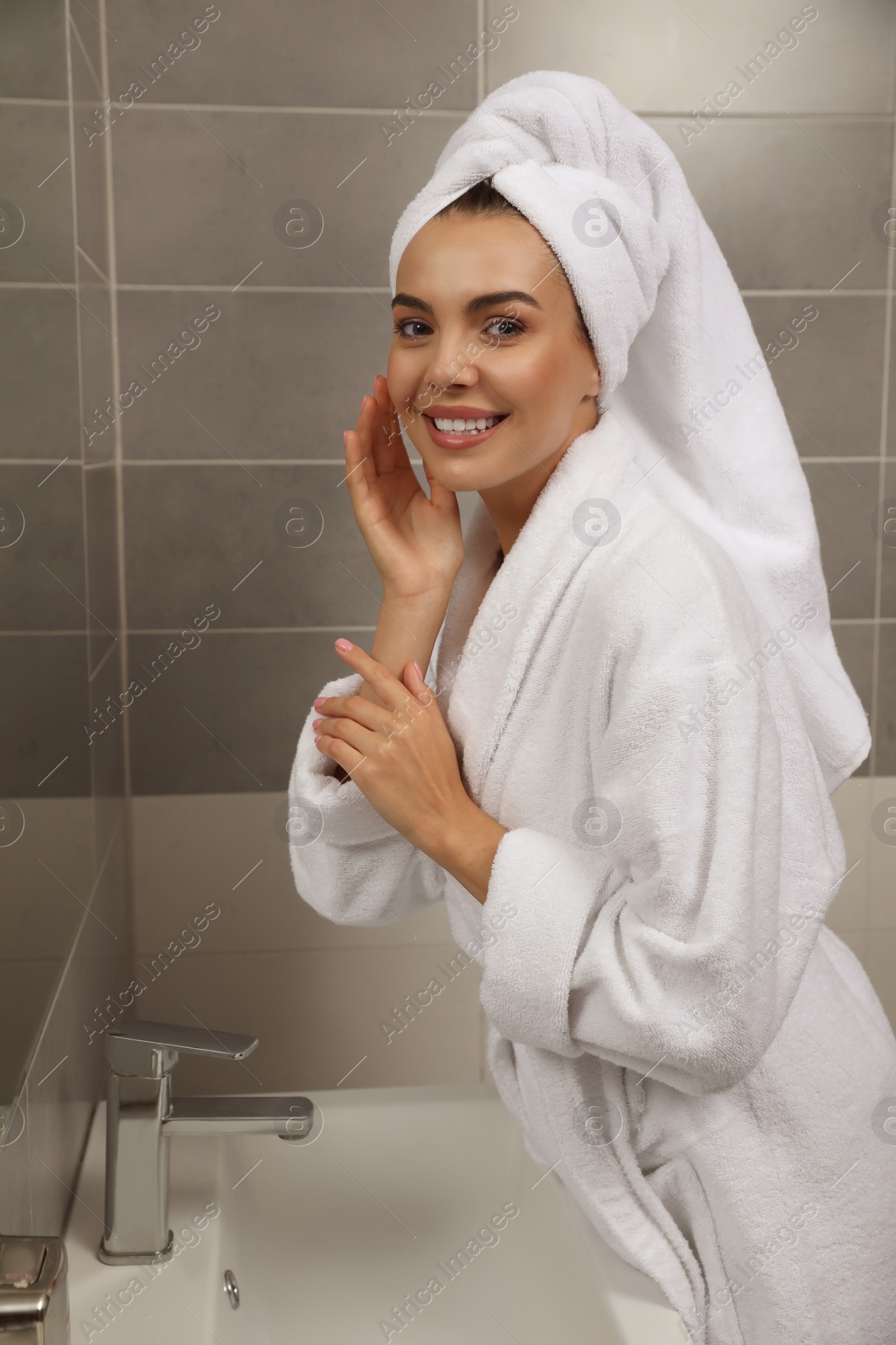 Photo of Beautiful young woman in bathrobe with towel on head near mirror at home