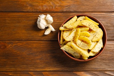 Photo of Bowl with tasty baked potato wedges, spices and garlic on wooden table, flat lay. Space for text