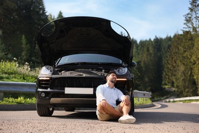Photo of Stressed man sitting on asphalt road near broken car outdoors