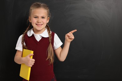 Photo of Happy schoolgirl with book pointing at something near blackboard, space for text