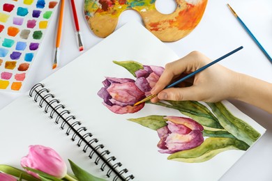 Photo of Woman painting tulips in sketchbook and supplies at white table, closeup