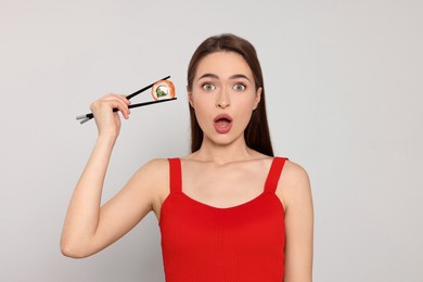 Emotional young woman holding sushi roll with chopsticks on light background