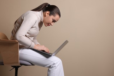 Young woman with poor posture using laptop while sitting on chair against beige background, space for text