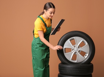 Female mechanic in uniform with car tires and clipboard on color background