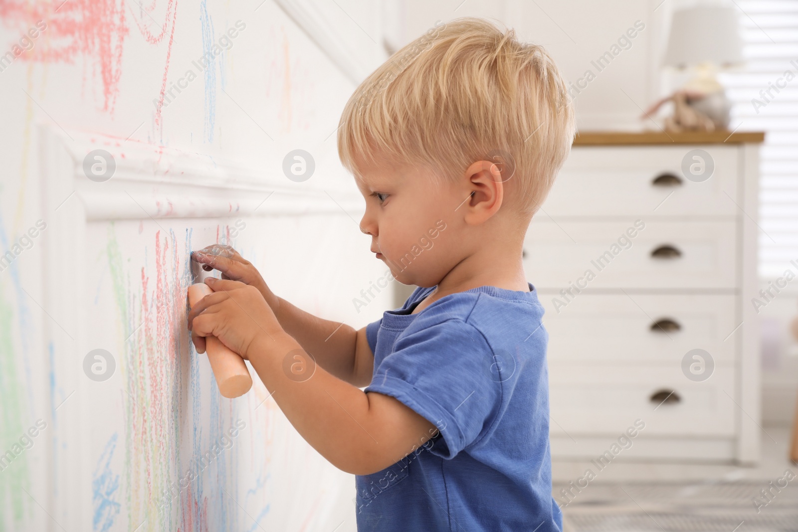 Photo of Mischievous little boy drawing with colorful chalk on white wall at home