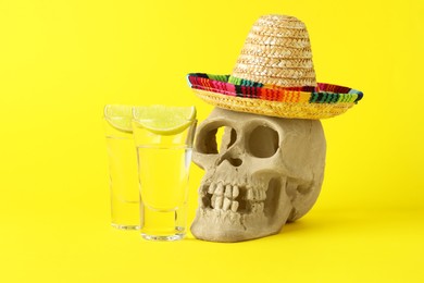 Photo of Human scull with Mexican sombrero hat, tequila in glasses and lime on yellow background