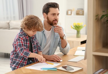 Photo of Man working remotely at home. Father using computer while his son drawing at desk