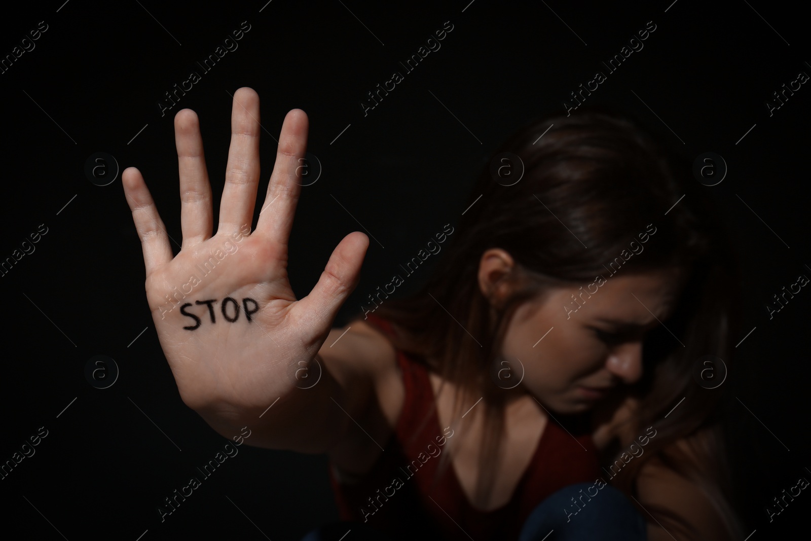 Photo of Crying young woman showing palm with word STOP against black background, focus on hand. Domestic violence concept