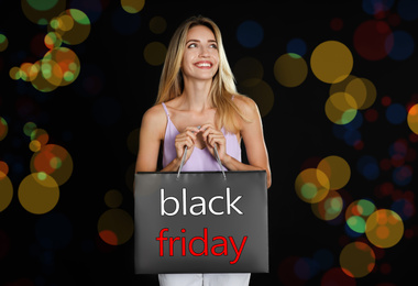 Black Friday sale. Happy young woman with shopping bag on dark background, bokeh effect