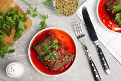 Photo of Plate of delicious stuffed grape leaves with tomato sauce, parsley and cutlery on light grey table, flat lay