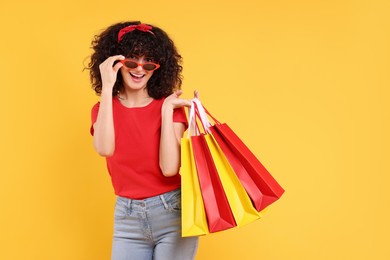 Happy young woman with shopping bags and stylish sunglasses on yellow background