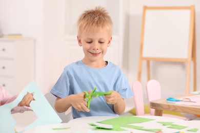 Photo of Happy little boy cutting green paper with scissors at desk in kindergarten. Playtime activities