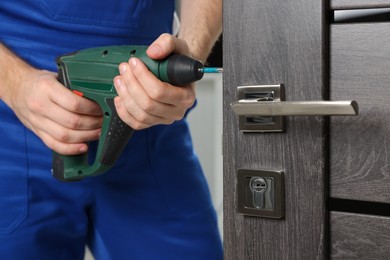 Photo of Worker in uniform with screw gun repairing door lock indoors, closeup