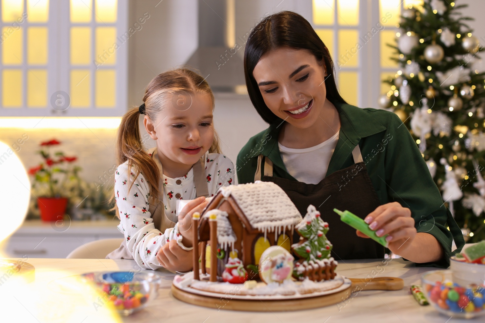 Photo of Mother and daughter decorating gingerbread house at table indoors