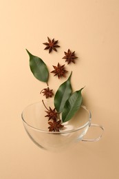 Anise stars and green leaves falling into glass cup on beige background, flat lay