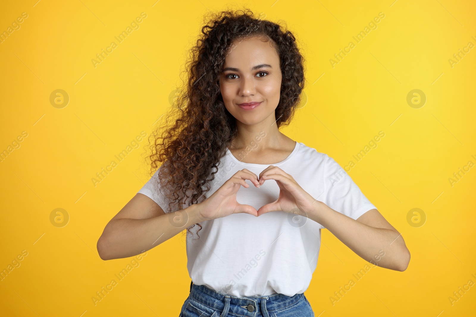 Photo of Happy young African-American woman making heart with hands on yellow background