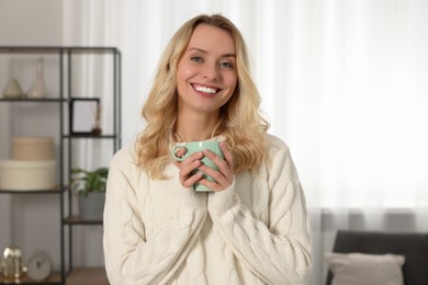 Photo of Happy woman in stylish warm sweater holding cup of drink at home