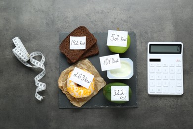 Photo of Calculator and food products with calorific value tags on dark grey table, flat lay. Weight loss concept