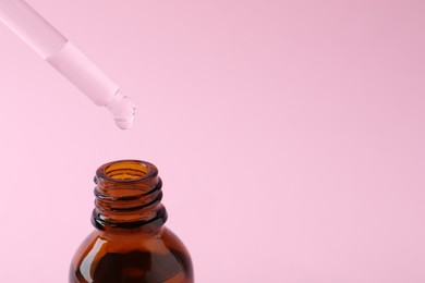 Dropping cosmetic oil from pipette into bottle on pink background, closeup. Space for text