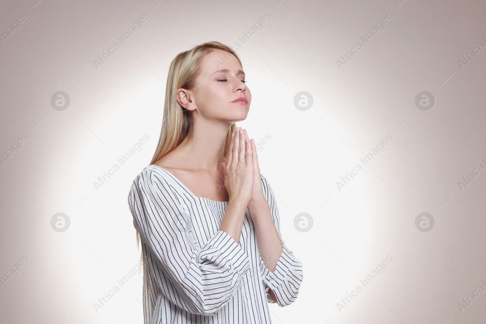 Photo of Religious young woman with clasped hands praying against light background. Space for text