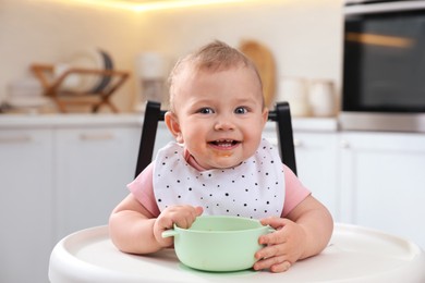 Photo of Cute little baby with bowl in high chair at kitchen