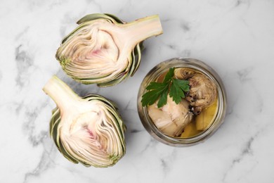 Jar of delicious artichokes pickled in olive oil and fresh vegetables on white marble table, flat lay