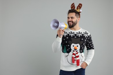 Young man in Christmas sweater and reindeer headband shouting in megaphone on grey background. Space for text