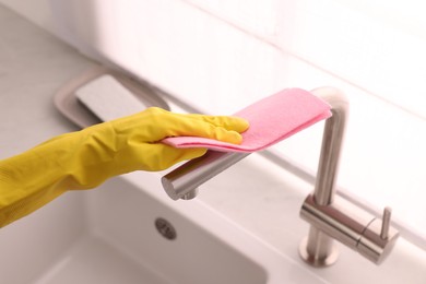 Photo of Woman in gloves cleaning faucet of kitchen sink with rag, closeup