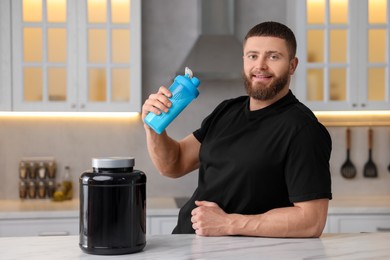 Young man with shaker of protein and powder at white marble table in kitchen