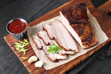 Photo of Pieces of baked pork belly served with sauce and parsley on black textured table, above view