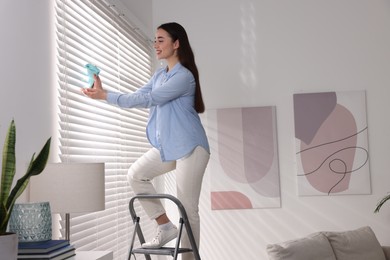 Woman on metal ladder wiping blinds at home