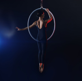 Young woman performing acrobatic element on aerial ring against dark background