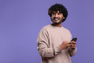 Handsome smiling man using smartphone on violet background, space for text